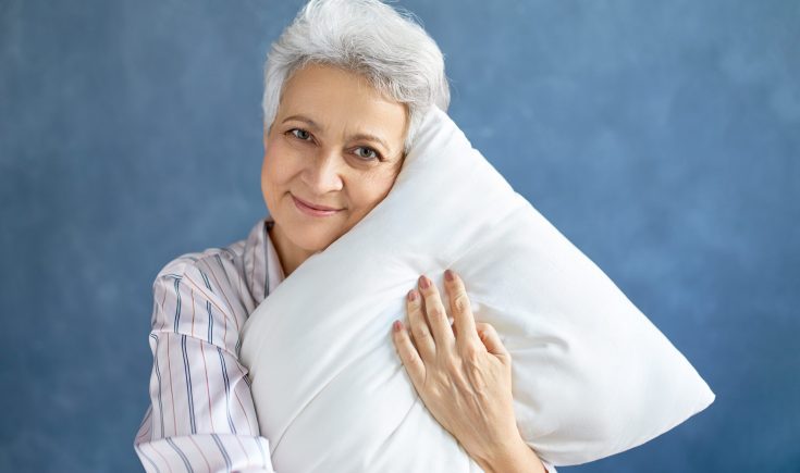 Bedding, relaxation and coziness concept. Isolated shot of cheerful charming middle aged woman with short gray hair hugging white pillow, lying awake, lazing in bed on weekend morning, smiling